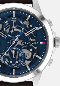 Montre à cadran bleu avec un boîtier en acier inoxydable, trois sous-cadrans et un bracelet en cuir marron. Nom de la marque "HILFIGER" affiché en évidence.
