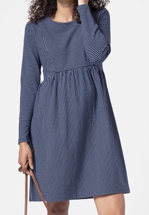 Woman wearing a navy blue dress with long sleeves and white vertical and horizontal stripes, holding a brown bag strap in one hand.