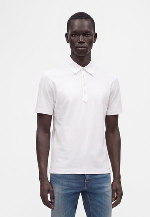 Homme portant un polo blanc uni à manches courtes et un jean bleu, debout devant un fond blanc, face à l'avant.
