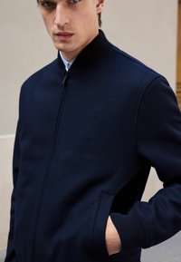 Tamsiai mėlynas bombonešio stilius striukė su stovinčiu apykaklę, priekiniais užtrauktukais ir šoninėmis kišenėmis; pagaminta iš tekstūruoto audinio su guminėmis rankovių rankovėmis.