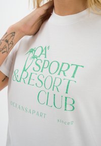 Biała koszulka z bawełny z zielonym nadrukiem graficznym „Sport & Resort Club” oraz wzorem palmy. Luźny krój z okrągłym dekoltem.