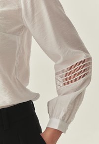 Chemise blanche texturée avec manches volumineuses. Détail notable avec découpe sur la manche, finition avec poignets ajustés et une ligne épurée.