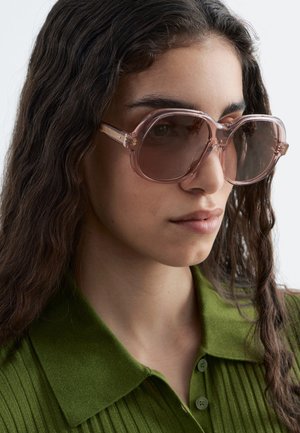 Vrouw met lang golvend haar, die oversized roze doorzichtige zonnebril en een groene geribde blouse met kraag draagt, kijkt iets opzij.