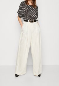 Schwarz-weiß gemusterte Bluse mit lässiger Passform, kombiniert mit hochtaillierten, weit geschnittenen cremefarbenen Hosen mit vertikalen Streifen und einem schwarzen Gürtel.