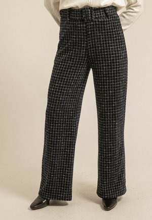 Pantalon large à carreaux noir et blanc avec passants de ceinture, porté avec des chaussures noires pointues et un haut beige clair rentré.