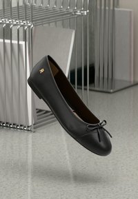 Ballerines en cuir noir avec un petit nœud et un logo doré, posées près d'un porte-revues en métal chromé sur un sol gris clair.