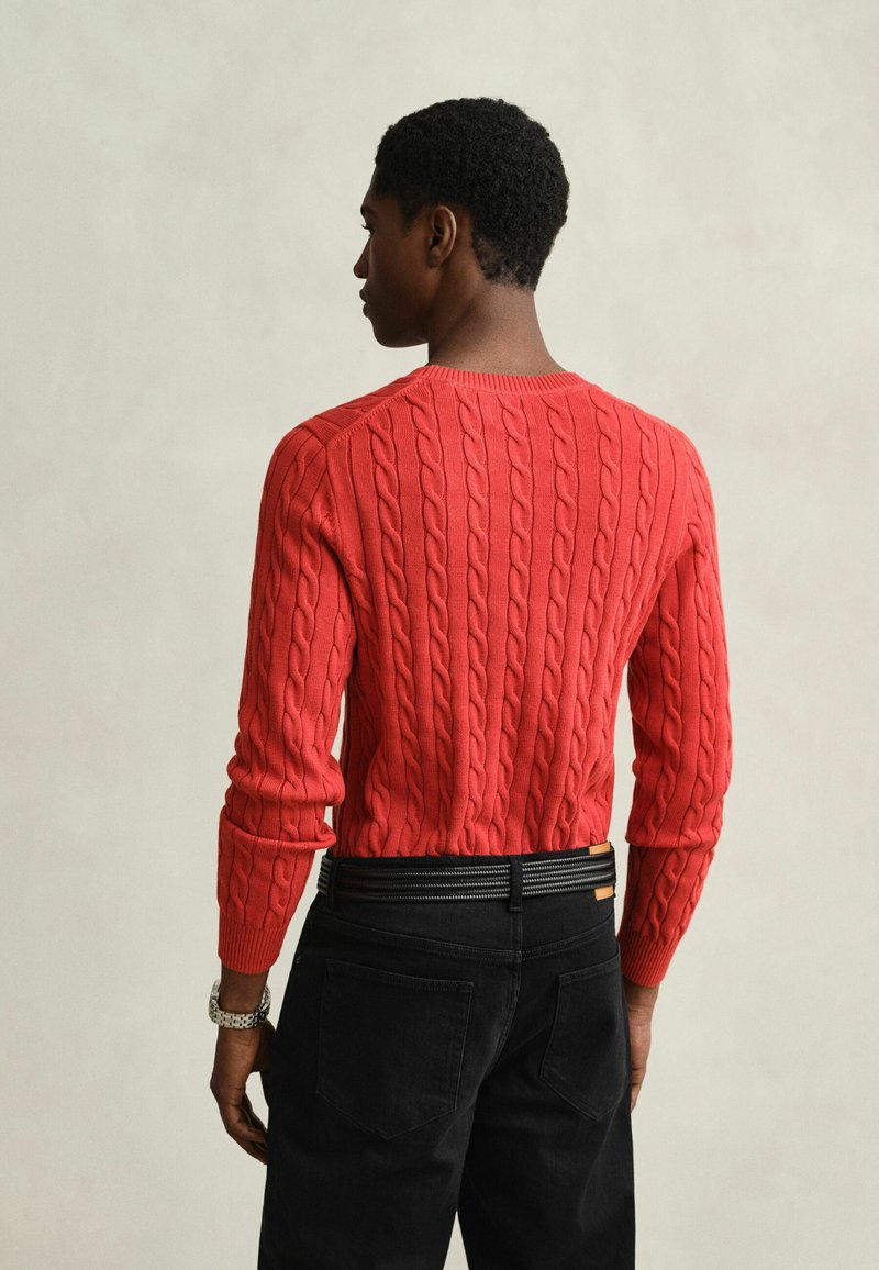 Man wearing a red cable-knit sweater and black pants, shown from the back against a plain light background.