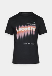 Črna bombažna majica s kratkimi rokavi, ki prikazuje gradientni logo "LACOSTE" v oranžni in roza barvi, z belim besedilom in zelenim krokodilom na prsih.