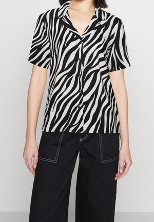 Kobieta ubrana w czarno-białą, zebra-pasiastą koszulę z krótkim rękawem na guziki, zestawioną z czarnymi spodniami z białym przeszyciem.