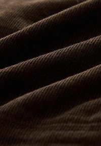 Bruin corduroy stof met zichtbare ribbels, zachte textuur en een lichte glans, die plooien en diepte in het materiaal toont.