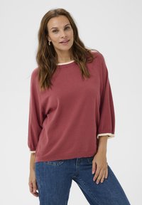 Woman with long brown hair wearing a loose red sweater with white trim and blue jeans, standing against a plain light background.