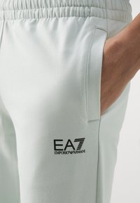 Detailní záběr na světle zbarvené tepláky s elastickým pasem, boční kapsou a černým logem EA7 Emporio Armani; ruka částečně v kapse.