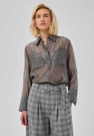 Vrouw met blond haar draagt een doorschijnend grijs overhemd met knopen en opgestikte zakken, en een grijs geruite broek met hoge taille en riem, staand tegen een effen achtergrond.