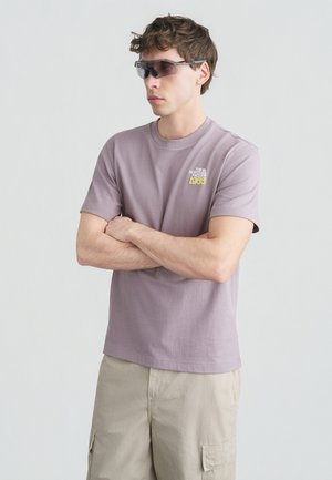 Νεαρός άντρας φορώντας μοβ μπλουζάκι The North Face και μπεζ cargo σορτς, με τα χέρια σταυρωμένα και φορώντας αθλητικά γυαλιά ηλίου με σκουρόχρωμους φακούς.