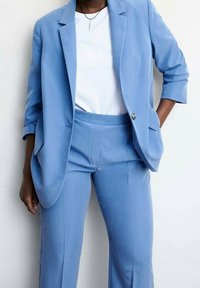 Costume bleu clair avec un blazer ajusté et un pantalon droit, associé à une chemise blanche. Tissu lisse avec des détails de design minimalistes.