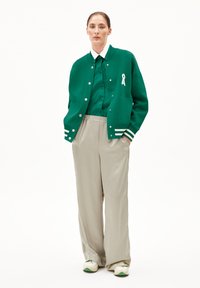 Veste varsity verte avec des rayures blanches et un détail en ruban, assortie à une chemise verte à boutons et un pantalon léger beige ample.