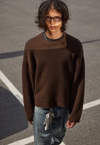 Jeune personne avec des cheveux bouclés, portant des lunettes, un pull brun, un jean bleu avec des clés et un tissu suspendus, se tenant à l'extérieur sur un sol pavé.