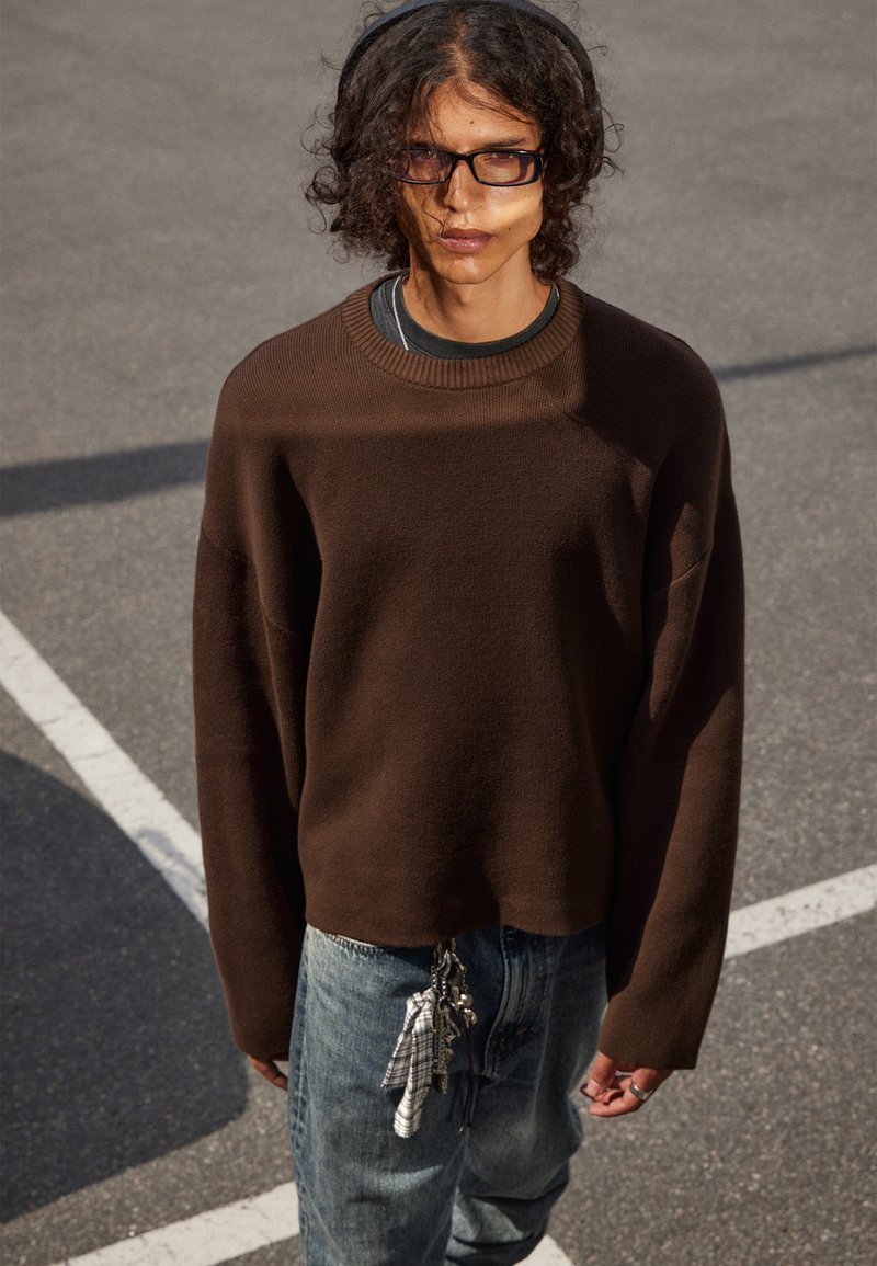 Jeune personne avec des cheveux bouclés, portant des lunettes, un pull brun, un jean bleu avec des clés et un tissu suspendus, se tenant à l'extérieur sur un sol pavé.