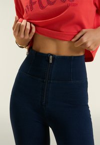 Jeans de cintura alta, de color azul oscuro, con cierre frontal de cremallera y textura de denim. Combinados con una blusa corta roja con adornos de piedras brillantes.
