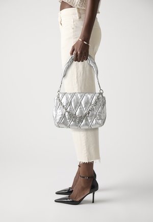 Silver quilted handbag with chain detail, held by a person wearing cream-colored pants and black pointed heels.