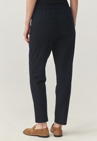 Pantaloni tapered bleu marin cu talie elastică, două buzunare la spate și o textură netedă. Purtați cu pantofi slip-on de culoare maro deschis.