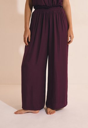 Personne portant un pantalon violet ample, taille haute, jambes larges, debout pieds nus sur un sol clair contre un mur beige.