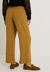 Femme portant un pantalon large moutarde, des mocassins en daim marron et un haut en dentelle noir à manches longues, se tenant devant un fond blanc uni.