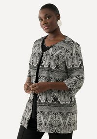 Giacca con motivo paisley in bianco e nero, maniche a tre quarti, scollatura rotonda e chiusura con bottoni sul davanti, indossata sopra un top nero.