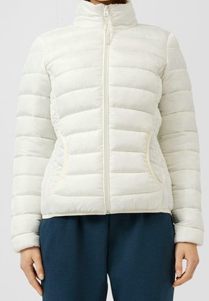 Personne portant une doudoune blanche matelassée avec des manches longues, une fermeture éclair devant et deux poches latérales, associée à un pantalon bleu foncé.