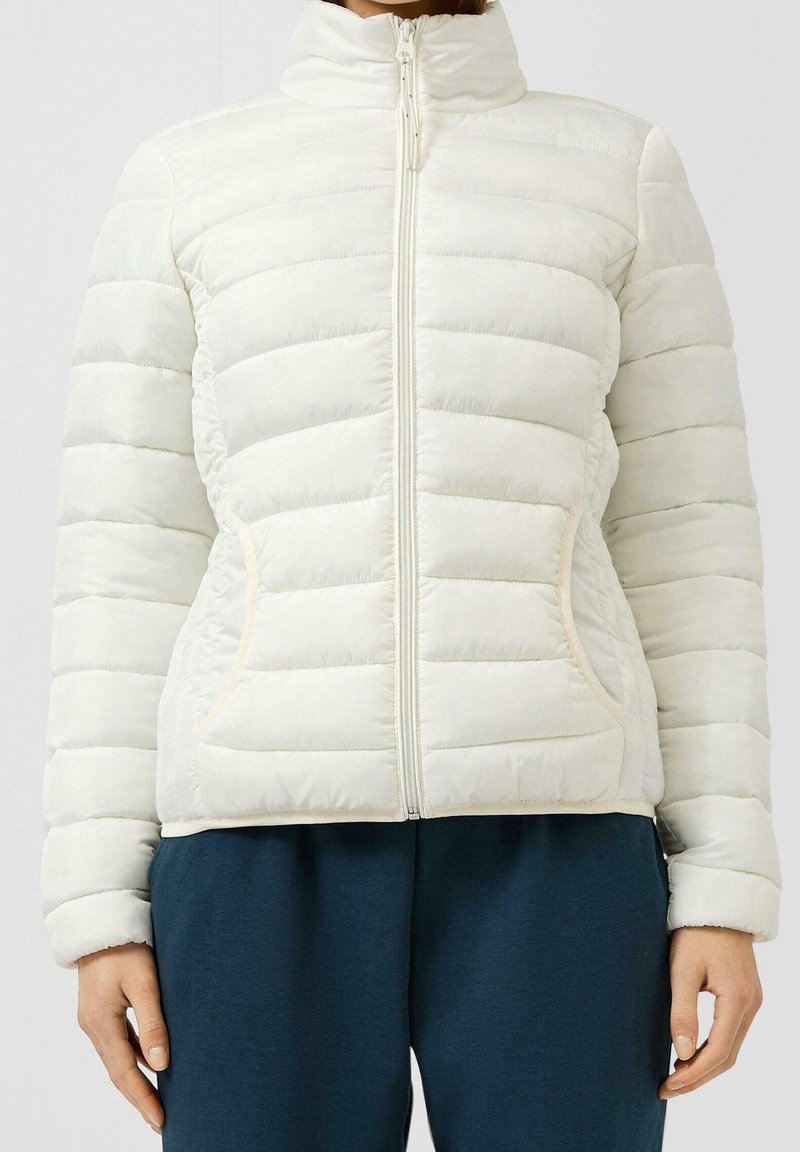 Personne portant une doudoune blanche matelassée avec des manches longues, une fermeture éclair devant et deux poches latérales, associée à un pantalon bleu foncé.