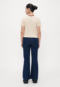 Beige short-sleeve t-shirt with a maroon neckline, paired with dark denim flared jeans and black platform shoes. Tattoos visible on arms.