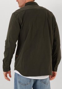 Man in een donkergroene, lange mouwen overhemd met knopen over een wit shirt, gecombineerd met lichtblauwe jeans, gezien vanuit de achterkant.