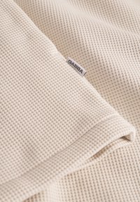 Vahvelikujuline kangas heledas kreemivärvis, millel on nähtav brändi silt. Pehme tekstuur ja voltidega äärte detail.