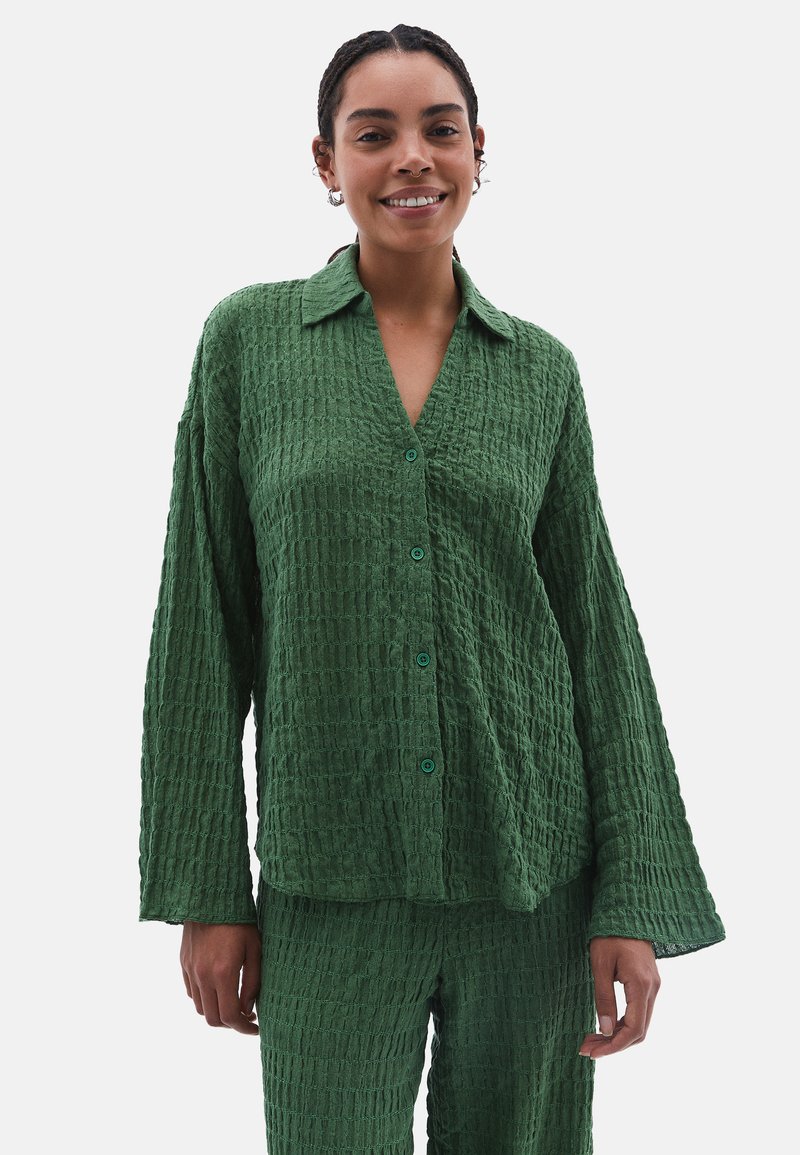 Camicia verde strutturata con colletto, maniche lunghe e chiusura con bottoni. Il tessuto presenta un motivo stropicciato, abbinato a pantaloni coordinati.