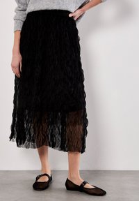 Gonna nera a strati realizzata in tessuto testurizzato, caratterizzata da un design ondulato e da un sovrapposto traslucido. Abbinata a ballerine nere con una semplice fascia.