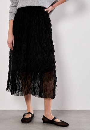 Schwarze, schichtweise Rock aus strukturiertem Stoff, mit einem wellenförmigen Design und einem durchsichtigen Überzug. Kombiniert mit schwarzen Ballerinas mit einem einfachen Riemen.