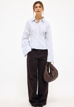 Kobieta w białej koszuli w prążki z szerokimi mankietami, czarnych spodni z szerokimi nogawkami, trzymająca brązową plecioną torebkę.