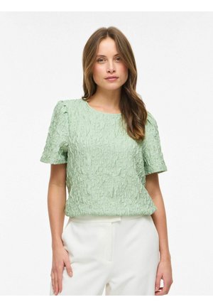 Femme aux cheveux bruns longs portant un haut vert clair texturé à manches courtes et un pantalon taille haute blanc, debout devant un fond uni.