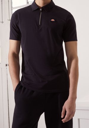 Homme portant un polo noir manches courtes avec une fermeture éclair au quart et un petit logo coloré sur la poitrine gauche, mains dans les poches d’un pantalon noir.