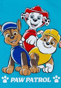 Grafica di Paw Patrol su una maglietta turchese che presenta tre cani dei cartoni animati: cane marrone in un gilet blu, cane bianco in rosso, cane marrone e bianco in giallo.