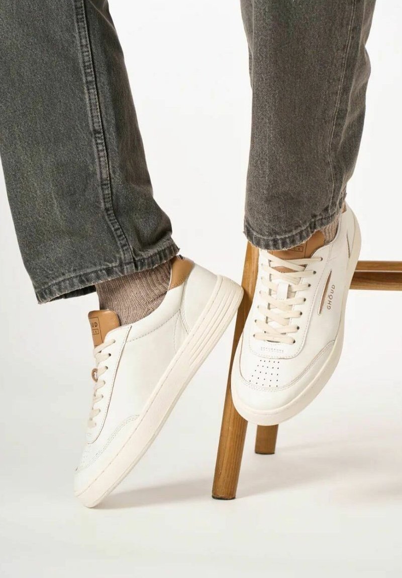 Sneakers bianchi in pelle con accenti beige, punta rotonda, dettagli perforati e lacci color crema. Indossati con jeans grigi e calze a coste.