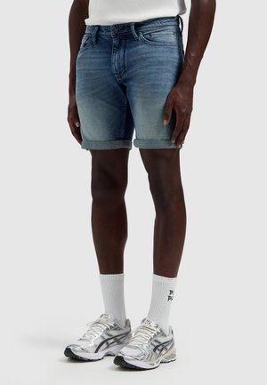 Man draagt opgerolde blauwe denimshorts, witte sportsokken met tekst en zilveren hardloopschoenen, staand voor een effen achtergrond.