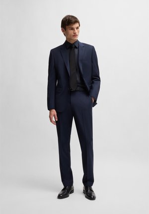 Marineblauw pak met een gestructureerd ontwerp, bestaande uit een enkelvoudig gesloten jack, bijpassende broek, zwart overhemd en stropdas, afgewerkt met zwarte schoenen.
