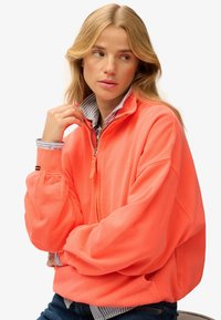 Pomarańczowy polar zapinany na zamek z wysokim kołnierzem, opadającymi ramionami i prążkowanymi detalami. Nosi się go na niebieskiej i białej koszuli w paski.