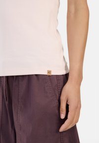 Light pink cotton tank top with side seams, featuring a small tan leather logo tag at the hem, paired with dark purple drawstring shorts.