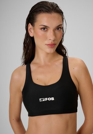 Kobieta z mokrymi włosami, ubrana w czarny sportowy biustonosz z logo "4F FOB", patrząca na wprost na jednolite szare tło.