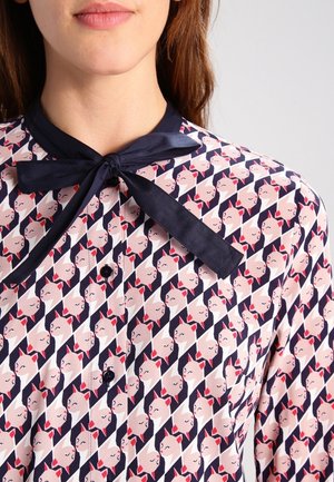 Kobieta w koszuli zapinanej na guziki z granatowym kołnierzykiem i kokardą, z powtarzającym się wzorem stylizowanych twarzy z czerwonymi kokardkami na różowym i granatowym tle.