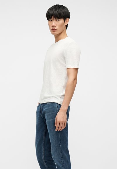 Modèle masculin debout de profil portant un t-shirt blanc uni à manches courtes et un jean en denim bleu sur un fond clair uni.