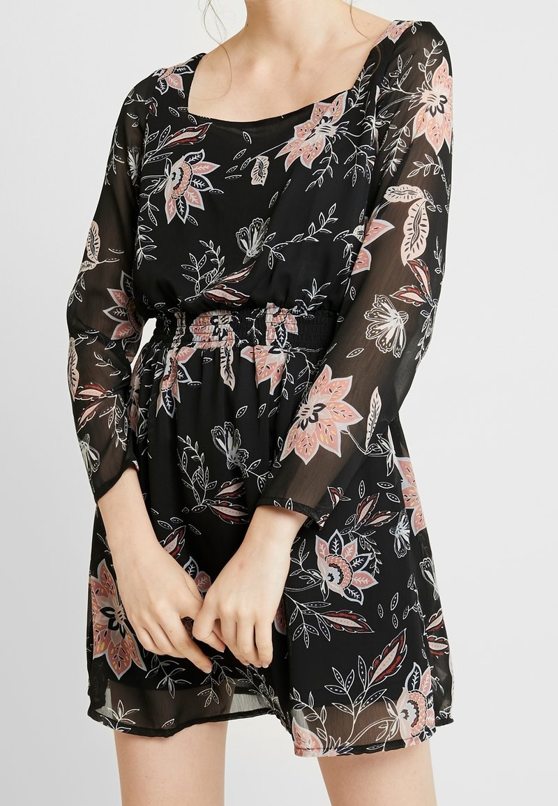 Robe noire à fleurs avec de longues manches transparentes. Dotée d'une taille smockée et d'un décolleté carré. Motifs floraux en rose et blanc.