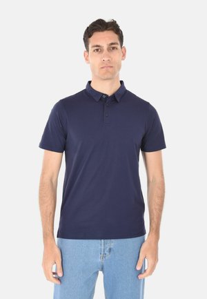 Uomo in piedi che indossa una polo a maniche corte blu navy e jeans azzurri chiari su uno sfondo bianco uniforme.